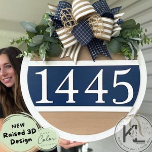 May include: A round wooden address sign with the number 1445 in white, set against a navy blue background. The sign features a white border, a natural wood grain base, and is adorned with a decorative bow and greenery. The sign reads "NEW Raised 3D Design MORE Color OPTIONS".