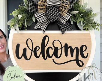 May include: A round wooden door sign featuring a light wood-toned center panel with the word Welcome in black script. The sign is accented with a large black and white patterned bow and green eucalyptus foliage at the top of the circular frame.