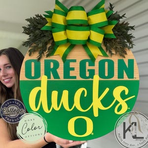 May include: A round wooden door sign with the words "OREGON ducks" in green and yellow. The sign has a green and wood grain background, a yellow and green bow, and greenery. The sign is a genuine college product.