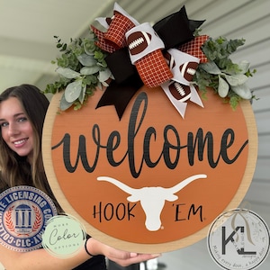 May include: Round wooden welcome sign with an orange center and a light wood border. The sign features the word "welcome" in black script, the phrase "Hook 'Em," and a white longhorn logo. It's decorated with a bow featuring footballs and greenery.