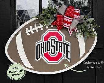 May include: A brown and white football-shaped door hanger with a raised 3D design. The football has the Ohio State logo in red, gray, and white. A red and white bow with gray accents is attached to the top of the football.