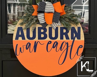 May include: A round wooden door hanger with an orange and wood background. The words "Auburn War Eagle" are written in navy blue lettering. A navy blue and white striped bow with orange polka dots is attached to the top of the hanger.
