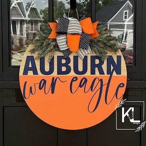 May include: A round wooden door hanger with an orange and wood background. The words "Auburn War Eagle" are written in navy blue lettering. A navy blue and white striped bow with orange polka dots is attached to the top of the hanger.