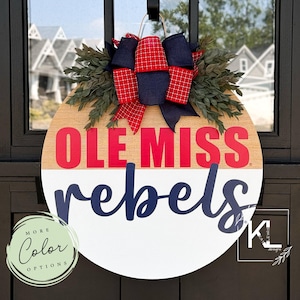 May include: A round wooden door hanger with a red and white design. The top half of the hanger is wood with the words "Ole Miss" in red. The bottom half is white with the word "rebels" in blue. A red and blue bow with white polka dots is attached to the top of the hanger.