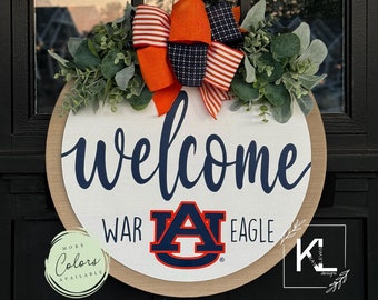 May include: A round wooden door hanger featuring the words "welcome", "war", "eagle", and the Auburn University logo. The door hanger is adorned with a red, white and blue bow and greenery.