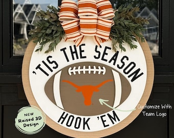 May include: A white and brown round door hanger with the text "'Tis the Season" and a football with a Longhorn logo. The text "Hook 'Em" is below the football. The door hanger is decorated with a brown and orange striped bow and green leaves.