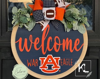 Può includere: Un appendiporta rotondo in legno con sfondo blu e le parole "welcome" in arancione. Il logo dell'Università di Auburn è al centro con le parole "War Eagle" su entrambi i lati. Un grande fiocco arancione e nero con palloni da calcio è in alto.