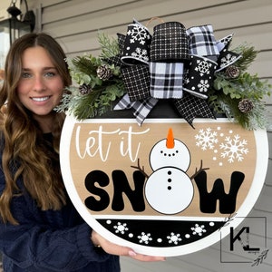 May include: A round wooden door hanger with a black and white plaid bow, evergreen sprigs, and pine cones. The door hanger features a snowman and the text "Let it Snow".