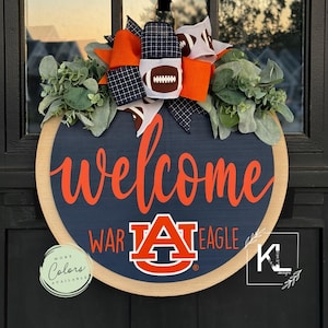 May include: A round wooden door hanger with a blue background and the words "welcome" in orange. The Auburn University logo is in the center with the words "War Eagle" on either side. A large orange and black bow with footballs is at the top.