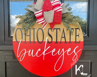 May include: A round wooden door hanger with a red and brown background. The words "Ohio State Buckeyes" are written in black lettering. A red and white striped bow with a gray bow is attached to the top of the hanger.