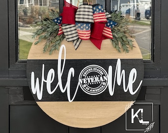 May include: A wooden welcome sign with black and white stripes and the word "welcome" written in white cursive. A red, white and blue bow with a plaid ribbon is attached to the top of the sign. A black and white sticker with the words "United States of America Veteran" is on the sign.