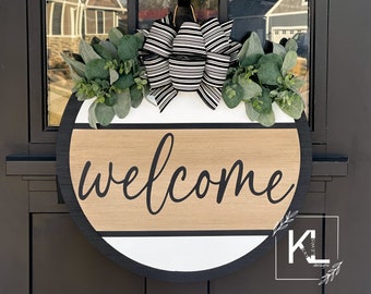 May include: A round wooden welcome sign with a black and white striped bow and greenery. The sign is painted white and brown with black lettering that says "welcome".