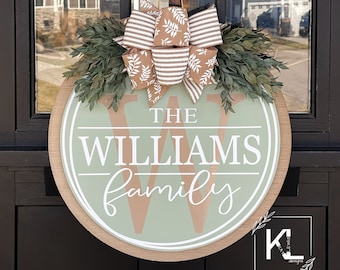 May include: A green and brown wooden door hanger with the text "THE WILLIAMS family" in white lettering. The hanger is decorated with a brown and white striped bow with a leafy design.