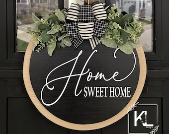 May include: A black and white wreath with a wooden frame and the text "Home Sweet Home" in white cursive lettering. The wreath is decorated with a black and white checkered bow and green eucalyptus leaves.
