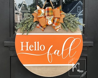 May include: A round wooden door hanger with the text "Hello fall" in white script on an orange background. The hanger is decorated with a plaid and floral bow and greenery.