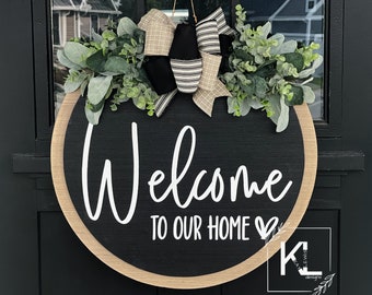 May include: Round wooden welcome sign with a black center and a light wood border. The sign reads "Welcome TO OUR HOME" in white script. It is decorated with greenery and bows and hangs on a black door.