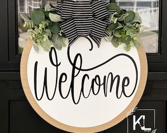 May include: A round wooden welcome sign with a black and white striped bow and greenery. The sign is hanging on a black door with the text "Welcome" in black cursive lettering. The sign is decorated with a small white logo that says "KL Designs".