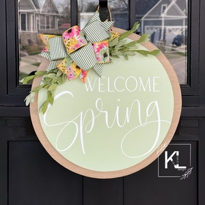 May include: A round wooden door hanger with a light green background and the text "Welcome Spring" in white cursive lettering. A green and white striped bow with floral print is attached to the top of the hanger.