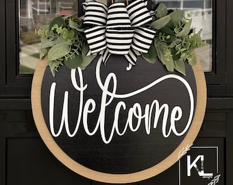 May include: Black and white striped bow with greenery on a round wooden door hanger with the word "Welcome" written in white cursive lettering.