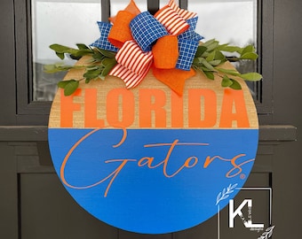 May include: A round wooden door hanger with the words "Florida Gators" in orange lettering on a blue background. The top of the hanger is decorated with a red, white, and blue bow and green leaves.