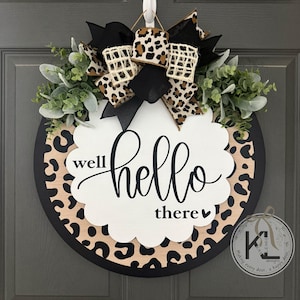 May include: Round wooden door wreath with a black border and a leopard print design. The wreath features a white scalloped shape with the words "well hello there" in black script. It is decorated with greenery and a bow.
