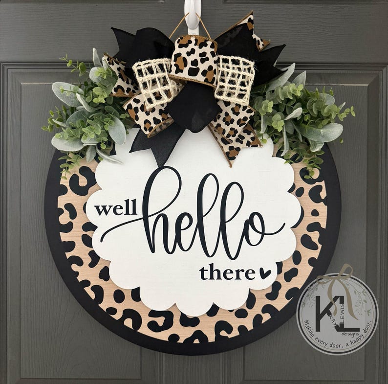 May include: Round wooden door wreath with a black border and a leopard print design. The wreath features a white scalloped shape with the words "well hello there" in black script. It is decorated with greenery and a bow.