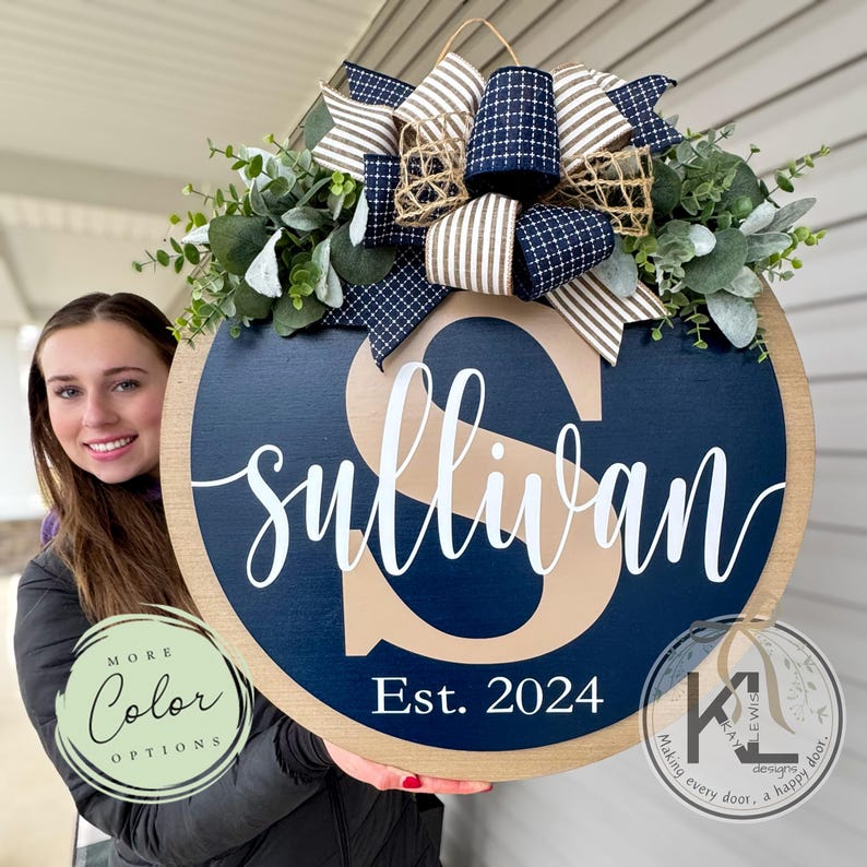 May include: A round wooden door hanger with a navy blue background and a large beige letter "S" in the center. The word "Sullivan" is written in white cursive script below the letter. The year "Est. 2024" is written in white below the name. The door hanger is decorated with a brown and white striped bow and greenery.
