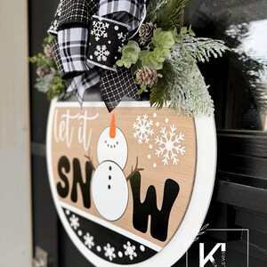 May include: A round wooden sign with the words "Let it Snow" in black and white lettering. A snowman is depicted in the center of the sign. The sign is decorated with a black and white plaid bow and a sprig of greenery.