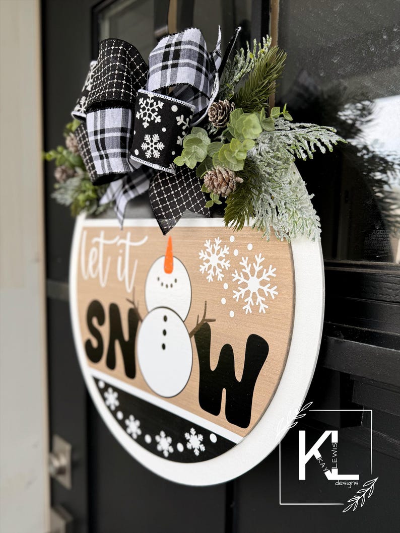 May include: A round wooden sign with the words "Let it Snow" in black and white lettering. A snowman is depicted in the center of the sign. The sign is decorated with a black and white plaid bow and a sprig of greenery.