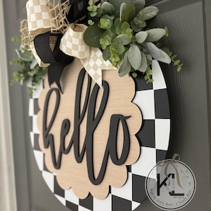 Op de afbeelding: Ronde houten deurhanger met een zwart-wit geblokt patroon en het woord "hello" in zwart schrift. De hanger is versierd met groen en een strik met een geblokt patroon.