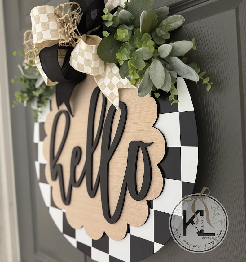 Op de afbeelding: Ronde houten deurhanger met een zwart-wit geblokt patroon en het woord "hello" in zwart schrift. De hanger is versierd met groen en een strik met een geblokt patroon.