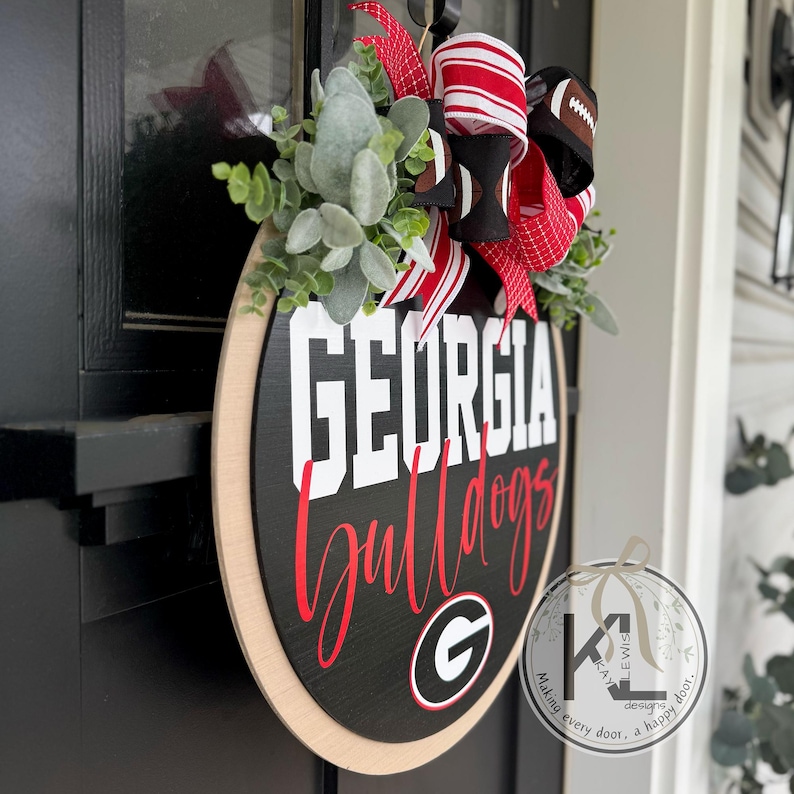 May include: A round wooden door hanger with the words "GEORGIA Bulldogs" in white and red lettering. The black background is accented with a light wood border, greenery, and a football-themed bow.