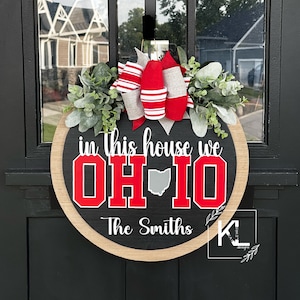 May include: A round wooden door sign with a black center and natural wood border. It features the white text in this house we, the red text OH-IO with a gray state silhouette, and the white text The Smiths. A red, white, and gray bow and greenery sit on top.