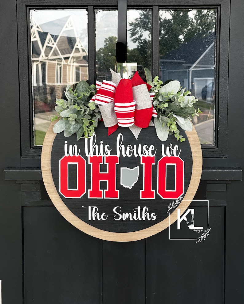 May include: A round wooden door sign with a black center and natural wood border. It features the white text in this house we, the red text OH-IO with a gray state silhouette, and the white text The Smiths. A red, white, and gray bow and greenery sit on top.