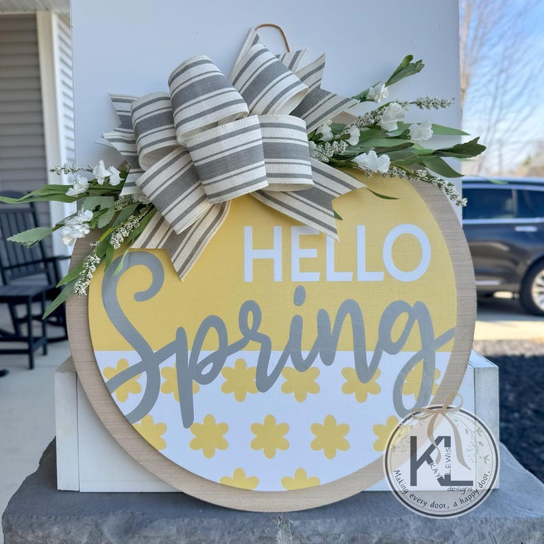 May include: A yellow and white round wooden door hanger with the text "Hello Spring" in gray lettering. The hanger is decorated with a gray and white striped bow and a sprig of white flowers.