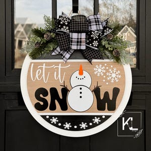 May include: A round wooden door hanger with the words "Let it Snow" in black and white lettering. A snowman with an orange carrot nose is in the center of the sign. The sign is decorated with a black and white plaid bow and a wreath of greenery.