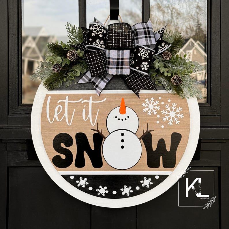 May include: A round wooden door hanger with the words "Let it Snow" in black and white lettering. A snowman with an orange carrot nose is in the center of the sign. The sign is decorated with a black and white plaid bow and a wreath of greenery.