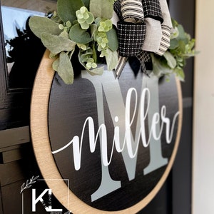 May include: Black and white wreath with a wooden round sign that says "Miller" with a gray letter M. The wreath is decorated with greenery and a black and white bow.