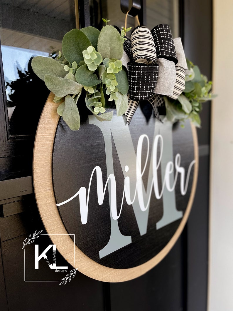 May include: Black and white wreath with a wooden round sign that says "Miller" with a gray letter M. The wreath is decorated with greenery and a black and white bow.