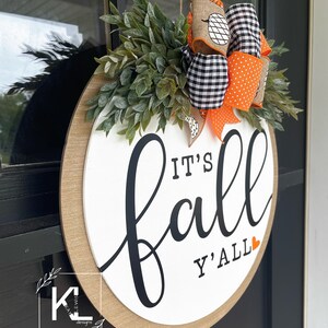 May include: A white round wooden sign with the text "It's Fall Y'all" in black script with a small orange heart. The sign is decorated with a black and white checkered ribbon with orange polka dots and a burlap ribbon with a pumpkin design. Green leafy sprigs surround the sign.