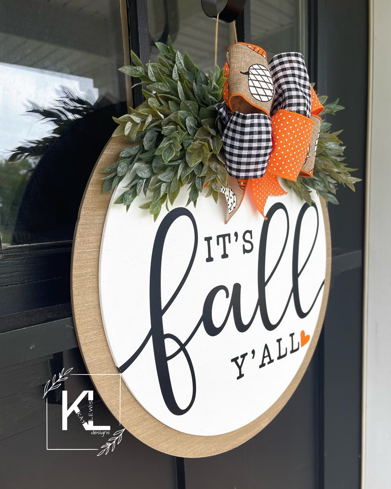 May include: A white round wooden sign with the text "It's Fall Y'all" in black script with a small orange heart. The sign is decorated with a black and white checkered ribbon with orange polka dots and a burlap ribbon with a pumpkin design. Green leafy sprigs surround the sign.