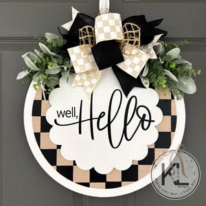 K&ouml;nnte beinhalten: Ein rundes T&uuml;rschild mit schwarz-beigem Schachbrettmuster und einem wei&szlig;en, gewellten Mittelst&uuml;ck, das den schwarzen Schriftzug well, HELLO tr&auml;gt. Das Schild ist mit einer gro&szlig;en Schleife aus schwarzen, karierten und Jute-B&auml;ndern sowie gr&uuml;nem Blattwerk verziert.