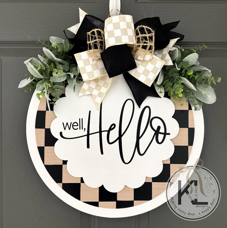 K&ouml;nnte beinhalten: Ein rundes T&uuml;rschild mit schwarz-beigem Schachbrettmuster und einem wei&szlig;en, gewellten Mittelst&uuml;ck, das den schwarzen Schriftzug well, HELLO tr&auml;gt. Das Schild ist mit einer gro&szlig;en Schleife aus schwarzen, karierten und Jute-B&auml;ndern sowie gr&uuml;nem Blattwerk verziert.