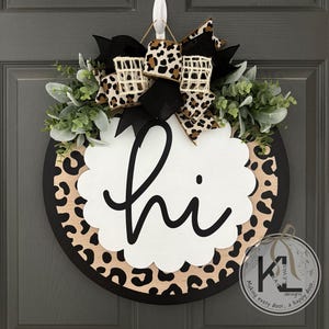 May include: Round wooden door wreath with a black border and a leopard print design. The wreath features a white scalloped center with the word "hi" in black script, accented with greenery and a leopard print bow.