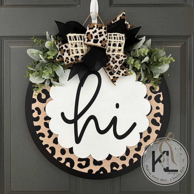 May include: Round wooden door wreath with a black border and a leopard print design. The wreath features a white scalloped center with the word "hi" in black script, accented with greenery and a leopard print bow.