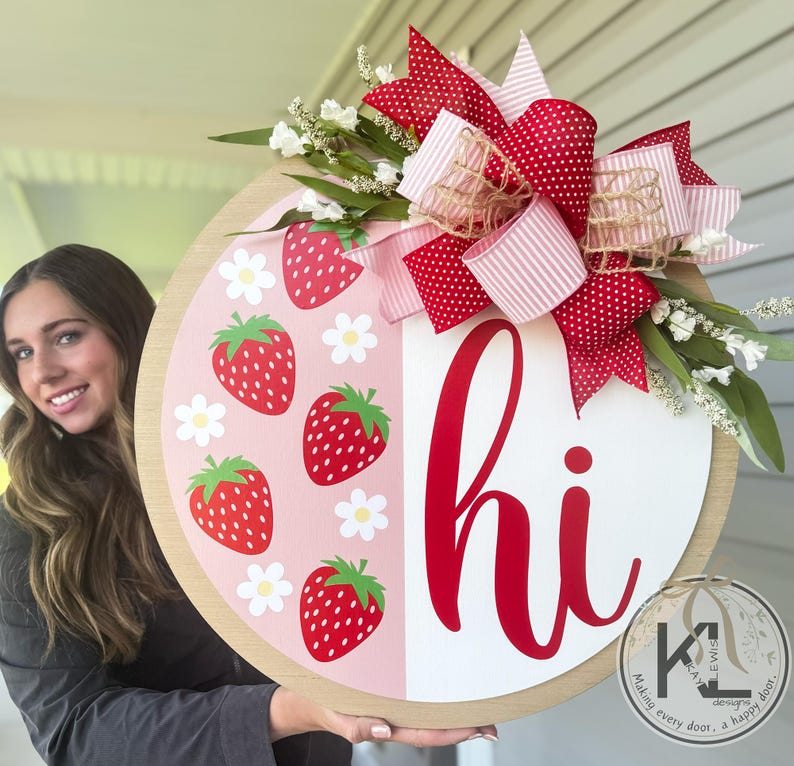 May include: A round wooden door hanger with a pink and white background. The left side features a pattern of red strawberries and white daisies. The right side is white with the word "hi" in red. A red and white polka dot bow with a pink and white striped ribbon is attached to the top.