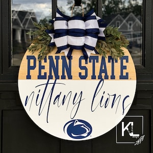 Puede incluir: Un colgador de puerta redondo de madera con fondo blanco y marr&oacute;n. La mitad superior es marr&oacute;n con el texto "Penn State" en letras azules. La mitad inferior es blanca con el texto "Nittany Lions" en escritura cursiva azul. Un lazo azul y blanco a rayas con hojas verdes est&aacute; unido a la parte superior del colgador. Un logotipo azul y blanco de Nittany Lion est&aacute; en el centro de la mitad inferior del colgador.