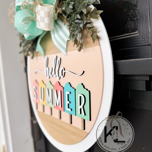 Pu&ograve; includere: Un appendiporta rotondo in legno con un bordo bianco. L'appendiporta ha uno sfondo rosa chiaro con le parole "hello summer" scritte in corsivo nero. La parola "summer" &egrave; composta da forme di ghiaccioli colorati.