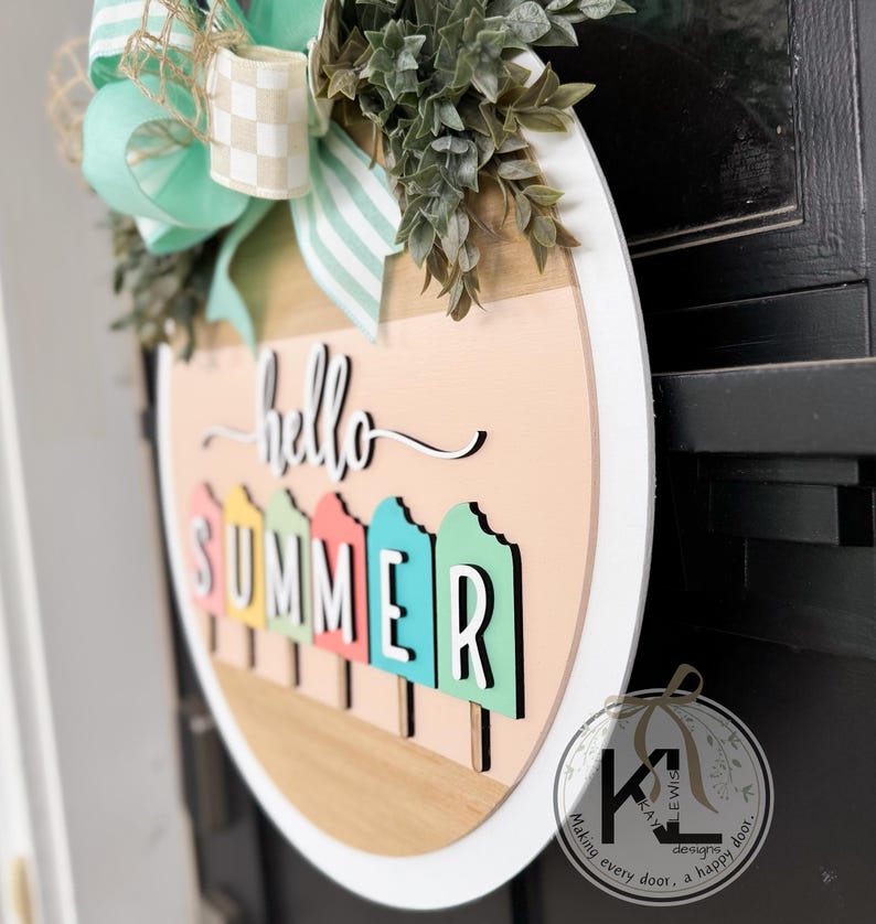 Pu&ograve; includere: Un appendiporta rotondo in legno con un bordo bianco. L'appendiporta ha uno sfondo rosa chiaro con le parole "hello summer" scritte in corsivo nero. La parola "summer" &egrave; composta da forme di ghiaccioli colorati.