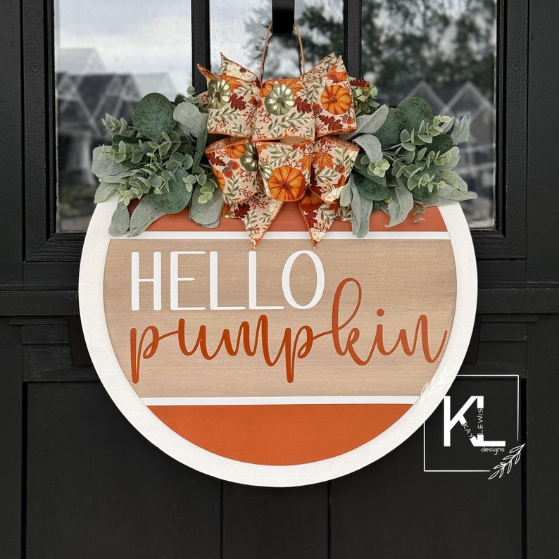 May include: A round wooden door hanger with a white background and orange and brown stripes. The words 'HELLO pumpkin' are written in white and orange lettering. The door hanger is decorated with a green and brown floral bow.